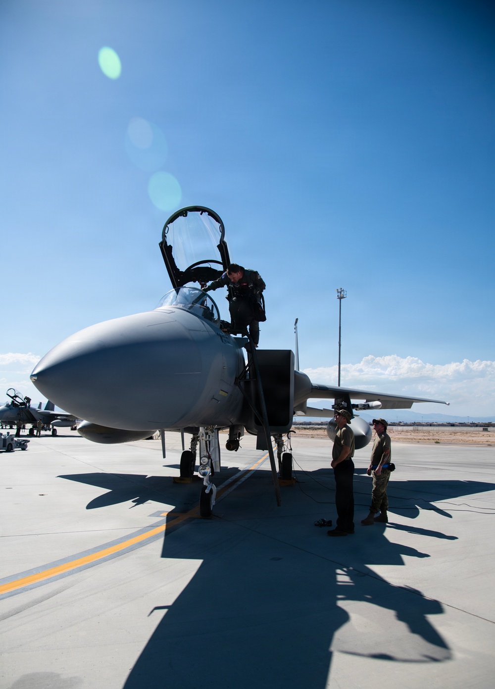 A military pilot is climbing out of the cockpit of an F-15 Eagle fighter jet on a flight line at what appears to be a desert air base. Two ground crew members stand near the aircraft, likely conducting post-flight procedures. The image is taken under bright, clear skies with lens flare visible from direct sunlight.