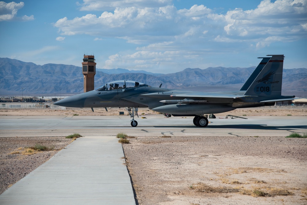 A U.S. military F-15 Eagle fighter jet with tail number '018' is taxiing or parked on a flight line at what appears to be a desert air base. The aircraft is in a gray paint scheme with colored tail markings and appears to have at least one crew member visible in the cockpit. An air traffic control tower and mountain range are visible in the background, consistent with a Southwestern U.S. desert installation.