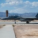 A U.S. military F-15 Eagle fighter jet with tail number '018' is taxiing or parked on a flight line at what appears to be a desert air base. The aircraft is in a gray paint scheme with colored tail markings and appears to have at least one crew member visible in the cockpit. An air traffic control tower and mountain range are visible in the background, consistent with a Southwestern U.S. desert installation.