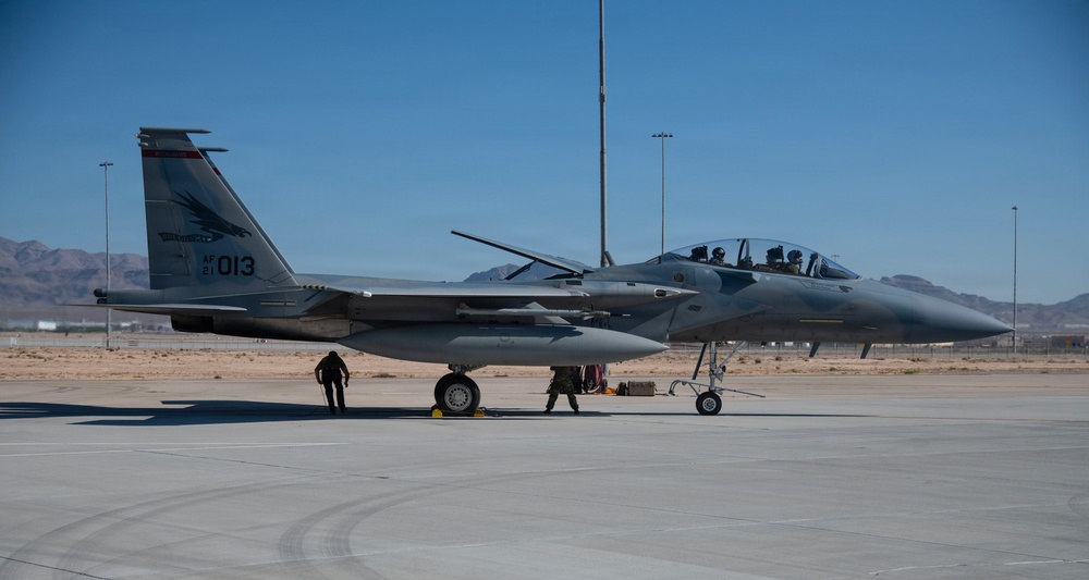 A U.S. Air Force F-15EX Eagle II fighter jet with tail number AF 21-013 is parked on a flight line, likely at Nellis Air Force Base in Nevada based on the desert terrain and mountain backdrop. Ground crew personnel are conducting pre-flight or post-flight checks around the aircraft, while at least one pilot is visible in the cockpit. The aircraft appears to be in an active operational configuration with weapons pylons and external stores visible.