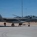 A U.S. Air Force F-15EX Eagle II fighter jet with tail number AF 21-013 is parked on a flight line, likely at Nellis Air Force Base in Nevada based on the desert terrain and mountain backdrop. Ground crew personnel are conducting pre-flight or post-flight checks around the aircraft, while at least one pilot is visible in the cockpit. The aircraft appears to be in an active operational configuration with weapons pylons and external stores visible.