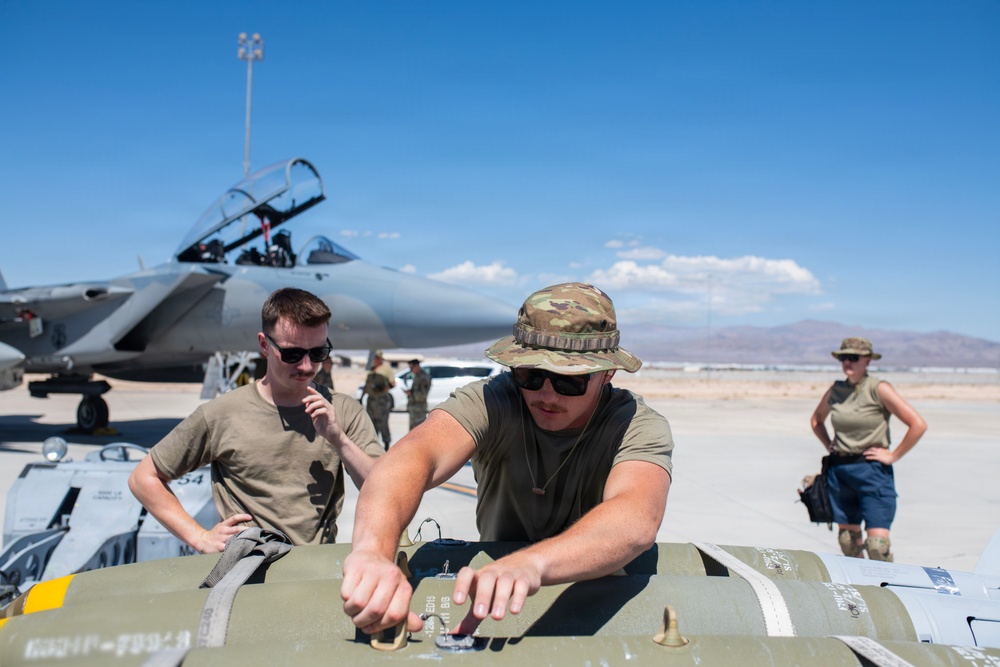 Military personnel are working on the flight line handling what appears to be munitions or ordnance on a cart, with a fighter jet (likely an F-15) visible in the background with its canopy open. The scene takes place on a desert airfield under clear blue skies, consistent with a southwestern U.S. military installation. At least three personnel are visible in the foreground, wearing olive drab t-shirts and tactical hats.