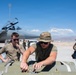 Military personnel are working on the flight line handling what appears to be munitions or ordnance on a cart, with a fighter jet (likely an F-15) visible in the background with its canopy open. The scene takes place on a desert airfield under clear blue skies, consistent with a southwestern U.S. military installation. At least three personnel are visible in the foreground, wearing olive drab t-shirts and tactical hats.