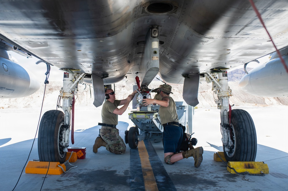 Two military maintenance personnel are working underneath a military aircraft, appearing to install or inspect a munition or equipment on the centerline hardpoint. The work is being conducted on a flight line in what appears to be an arid desert environment. Both individuals are in military-style attire and are kneeling beneath the fuselage using a munitions loading cart.