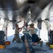 Two military maintenance personnel are working underneath a military aircraft, appearing to install or inspect a munition or equipment on the centerline hardpoint. The work is being conducted on a flight line in what appears to be an arid desert environment. Both individuals are in military-style attire and are kneeling beneath the fuselage using a munitions loading cart.