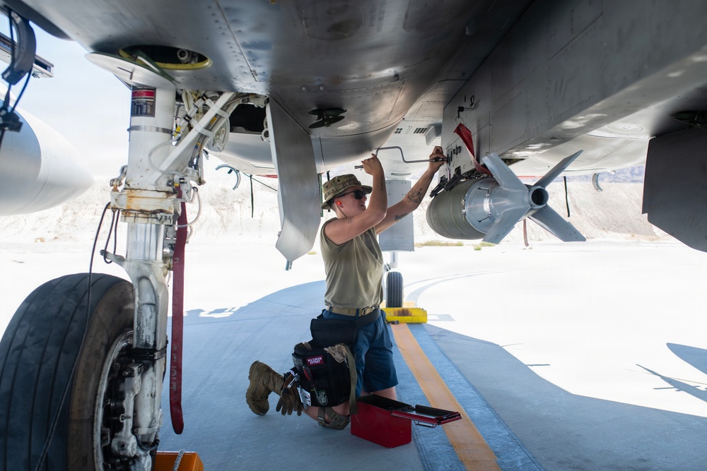 A military aircraft maintainer or weapons technician is kneeling beneath a military jet aircraft on a flight line, performing maintenance or weapons loading work. The individual is working on what appears to be an ordnance or munition mounted under the aircraft's wing or fuselage. The setting appears to be a desert air base based on the bright, arid environment visible in the background.