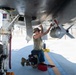 A military aircraft maintainer or weapons technician is kneeling beneath a military jet aircraft on a flight line, performing maintenance or weapons loading work. The individual is working on what appears to be an ordnance or munition mounted under the aircraft's wing or fuselage. The setting appears to be a desert air base based on the bright, arid environment visible in the background.