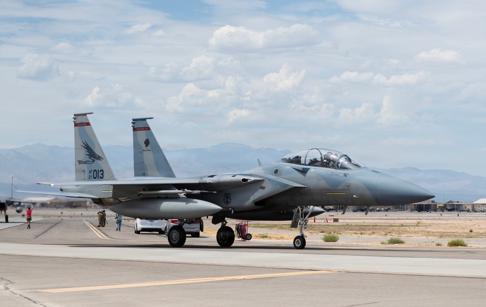 A U.S. Air Force F-15 Eagle fighter jet, tail number 013, is taxiing on a flight line at what appears to be a desert military airbase. The aircraft is in a two-seat configuration and displays unit markings on its twin tail fins. Ground personnel and support vehicles are visible nearby on the tarmac.