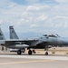 A U.S. Air Force F-15 Eagle fighter jet, tail number 013, is taxiing on a flight line at what appears to be a desert military airbase. The aircraft is in a two-seat configuration and displays unit markings on its twin tail fins. Ground personnel and support vehicles are visible nearby on the tarmac.