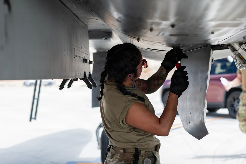 A military aircraft maintainer, appearing to be a female service member, is performing maintenance work on the underside of a military aircraft on a flight line or tarmac. She is using a hand tool and wearing black gloves, a tan/olive uniform, and has visible tattoos on her arm. A second person is partially visible in the background near a vehicle.