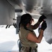 A military aircraft maintainer, appearing to be a female service member, is performing maintenance work on the underside of a military aircraft on a flight line or tarmac. She is using a hand tool and wearing black gloves, a tan/olive uniform, and has visible tattoos on her arm. A second person is partially visible in the background near a vehicle.