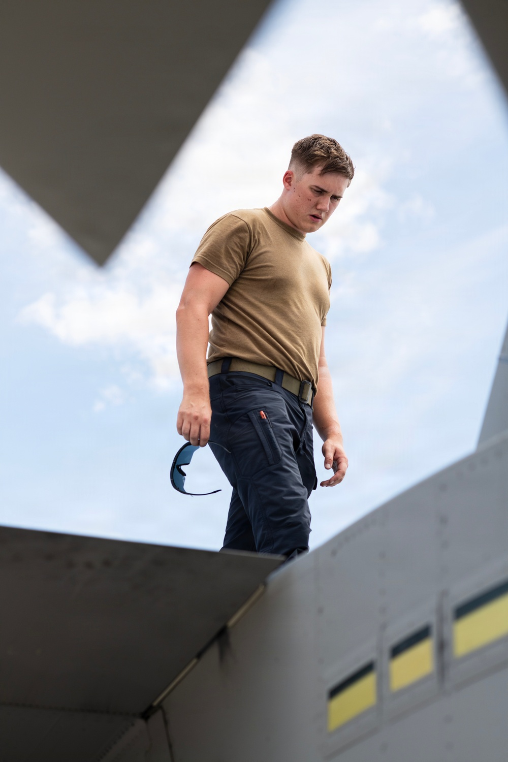 A young male in military-style attire is photographed from a low angle, standing on or near a large gray structure, likely an aircraft or military vehicle. He is wearing a tan/coyote brown t-shirt, dark navy tactical pants with a belt, and is holding a pair of blue-tinted protective eyewear in his left hand. The dramatic upward perspective frames him against a partly cloudy sky.