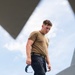 A young male in military-style attire is photographed from a low angle, standing on or near a large gray structure, likely an aircraft or military vehicle. He is wearing a tan/coyote brown t-shirt, dark navy tactical pants with a belt, and is holding a pair of blue-tinted protective eyewear in his left hand. The dramatic upward perspective frames him against a partly cloudy sky.