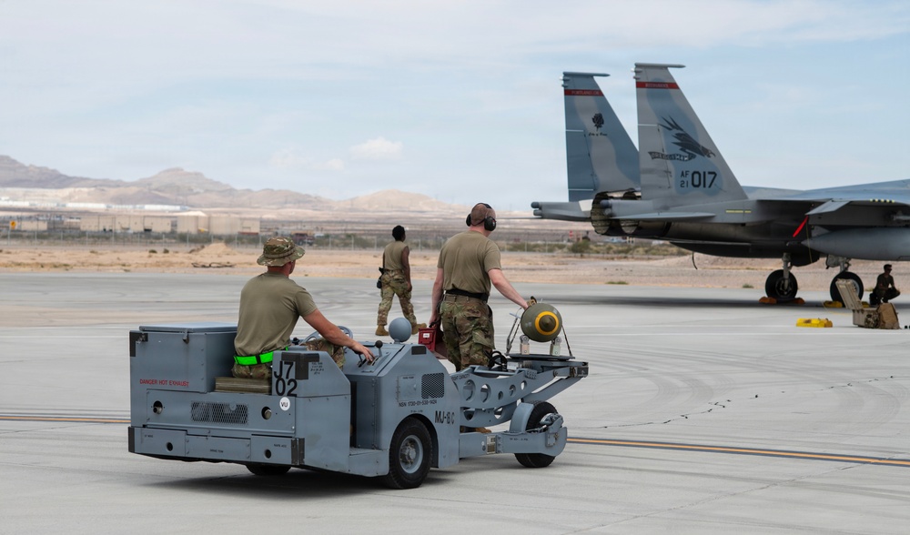 Military ground crew personnel are operating on an airfield tarmac, transporting what appears to be an unguided bomb or munition on an MJ-8C weapons loader cart toward an F-15 Eagle fighter jet. The aircraft tail number AF 21-017 is clearly visible, along with squadron markings. The setting appears to be a desert military airbase, likely in the southwestern United States.