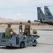 Military ground crew personnel are operating on an airfield tarmac, transporting what appears to be an unguided bomb or munition on an MJ-8C weapons loader cart toward an F-15 Eagle fighter jet. The aircraft tail number AF 21-017 is clearly visible, along with squadron markings. The setting appears to be a desert military airbase, likely in the southwestern United States.