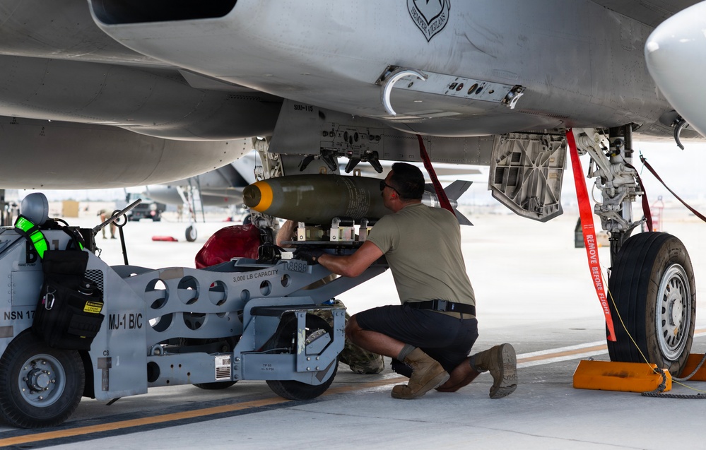 A military aircraft maintainer is loading or securing a munition onto the underside of a military jet aircraft on a flight line. The technician is using an MJ-1B/C weapons loading cart, rated at 3,000 lb capacity, to position what appears to be a bomb or guided munition onto a hardpoint beneath the aircraft. Safety equipment including red 'Remove Before Flight' streamers and wheel chocks are visible.