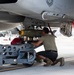 A military aircraft maintainer is loading or securing a munition onto the underside of a military jet aircraft on a flight line. The technician is using an MJ-1B/C weapons loading cart, rated at 3,000 lb capacity, to position what appears to be a bomb or guided munition onto a hardpoint beneath the aircraft. Safety equipment including red 'Remove Before Flight' streamers and wheel chocks are visible.