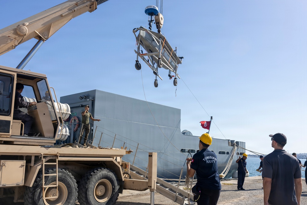 A military loading operation is taking place at a naval dock, where a crane mounted on a tan/desert-colored military vehicle is lifting what appears to be a folded stretcher or medical equipment. Personnel from the Tunisian Navy, identifiable by text on their uniforms and a red Tunisian flag visible on the vessel, are supervising the operation alongside other military personnel. A large gray naval vessel is moored at the dock in the background.