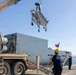 A military loading operation is taking place at a naval dock, where a crane mounted on a tan/desert-colored military vehicle is lifting what appears to be a folded stretcher or medical equipment. Personnel from the Tunisian Navy, identifiable by text on their uniforms and a red Tunisian flag visible on the vessel, are supervising the operation alongside other military personnel. A large gray naval vessel is moored at the dock in the background.