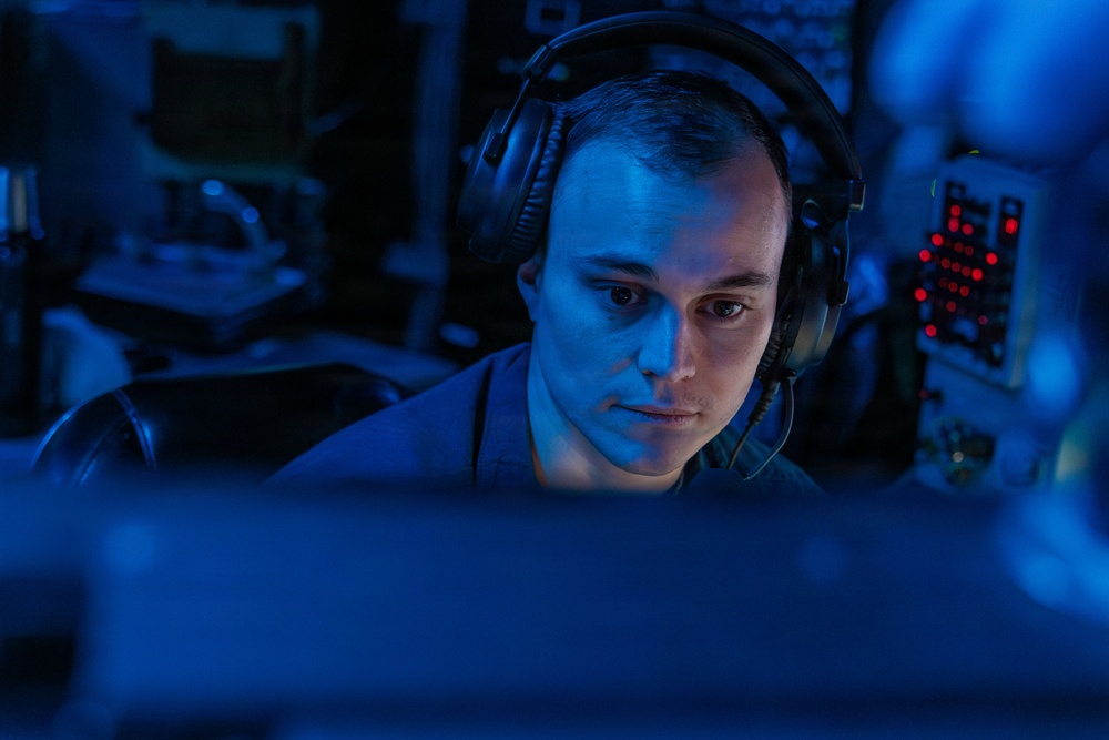 A young adult male wearing over-ear headphones is seated in a dimly lit environment illuminated predominantly by blue light, intently focused on a screen in the foreground. The background contains electronic equipment with visible red LED indicator lights, suggesting a technical or operational workspace. The overall atmosphere is dark and concentrated, consistent with a monitoring, gaming, or communications setting.