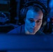 A young adult male wearing over-ear headphones is seated in a dimly lit environment illuminated predominantly by blue light, intently focused on a screen in the foreground. The background contains electronic equipment with visible red LED indicator lights, suggesting a technical or operational workspace. The overall atmosphere is dark and concentrated, consistent with a monitoring, gaming, or communications setting.