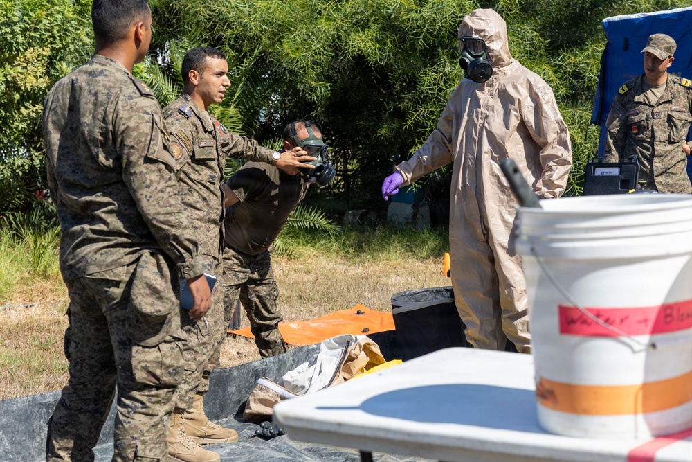 Military personnel are conducting what appears to be a CBRN (Chemical, Biological, Radiological, and Nuclear) training exercise outdoors. One individual is wearing a full hazmat suit with gas mask and purple nitrile gloves, while others in standard military camouflage uniforms observe and interact. Equipment including a white bucket labeled with red text, orange tarp, and detection devices are visible on a table.