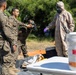 Military personnel are conducting what appears to be a CBRN (Chemical, Biological, Radiological, and Nuclear) training exercise outdoors. One individual is wearing a full hazmat suit with gas mask and purple nitrile gloves, while others in standard military camouflage uniforms observe and interact. Equipment including a white bucket labeled with red text, orange tarp, and detection devices are visible on a table.