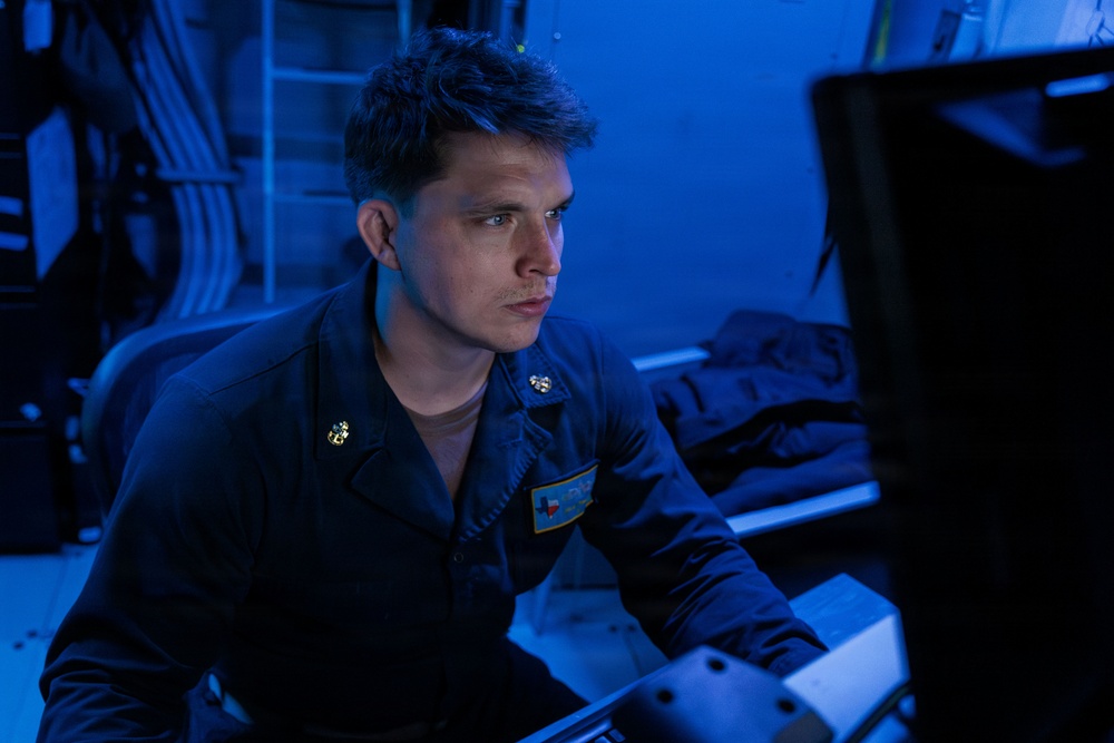 A young male military personnel, appearing to be a U.S. Navy sailor, is seated at a workstation in a dimly lit, blue-illuminated environment consistent with a naval vessel's combat information center or operations room. He is wearing a dark navy coverall uniform with gold insignia pins and a name/unit patch visible on the chest. His attention is directed toward a screen or display out of frame to the right.