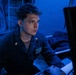 A young male military personnel, appearing to be a U.S. Navy sailor, is seated at a workstation in a dimly lit, blue-illuminated environment consistent with a naval vessel's combat information center or operations room. He is wearing a dark navy coverall uniform with gold insignia pins and a name/unit patch visible on the chest. His attention is directed toward a screen or display out of frame to the right.