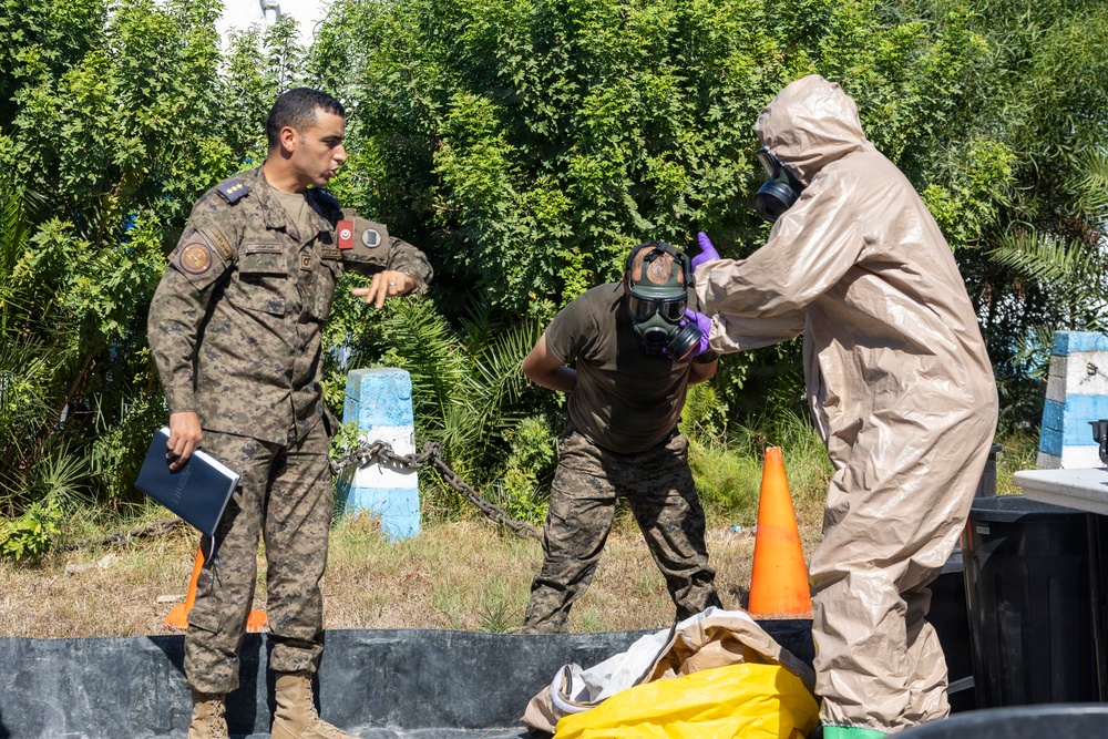 The image shows three military personnel engaged in what appears to be a CBRN (Chemical, Biological, Radiological, Nuclear) training exercise or decontamination drill. One person in full hazmat/protective suit with gas mask and purple gloves is assisting a second person wearing a gas mask and military fatigues, while a third uniformed officer observes and appears to be providing instruction. The setting is an outdoor area with vegetation in the background.