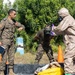 The image shows three military personnel engaged in what appears to be a CBRN (Chemical, Biological, Radiological, Nuclear) training exercise or decontamination drill. One person in full hazmat/protective suit with gas mask and purple gloves is assisting a second person wearing a gas mask and military fatigues, while a third uniformed officer observes and appears to be providing instruction. The setting is an outdoor area with vegetation in the background.