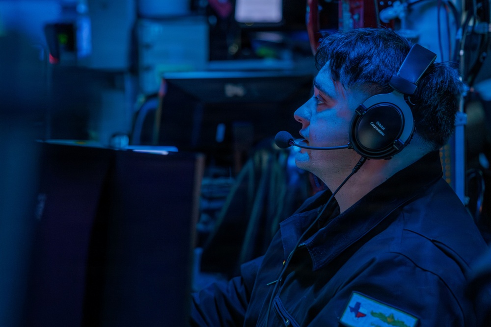 A male operator wearing a military-style uniform and communication headset with boom microphone is seated at a workstation in a dimly lit control room or combat information center. The environment is illuminated predominantly by blue ambient lighting, consistent with naval operations centers or similar tactical environments. Multiple monitors and electronic equipment are visible in the background.