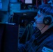 A male operator wearing a military-style uniform and communication headset with boom microphone is seated at a workstation in a dimly lit control room or combat information center. The environment is illuminated predominantly by blue ambient lighting, consistent with naval operations centers or similar tactical environments. Multiple monitors and electronic equipment are visible in the background.