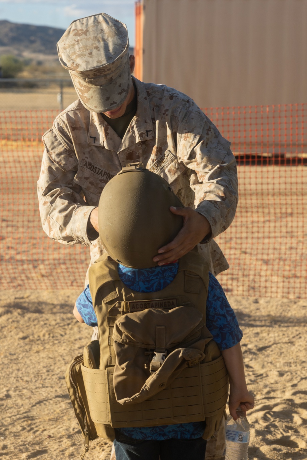 A U.S. Marine in desert MARPAT camouflage uniform is placing a military combat helmet onto a small child who is already wearing an oversized tactical vest/plate carrier. Both the Marine's uniform and the child's vest display the name tape 'ACOSTAPINEDA', suggesting a family relationship. The scene takes place outdoors in an arid, sandy environment consistent with a military base or training area.