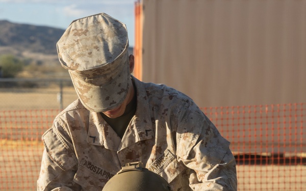 A U.S. Marine in desert MARPAT camouflage uniform is placing a military combat helmet onto a small child who is already wearing an oversized tactical vest/plate carrier. Both the Marine's uniform and the child's vest display the name tape 'ACOSTAPINEDA', suggesting a family relationship. The scene takes place outdoors in an arid, sandy environment consistent with a military base or training area.