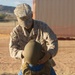 A U.S. Marine in desert MARPAT camouflage uniform is placing a military combat helmet onto a small child who is already wearing an oversized tactical vest/plate carrier. Both the Marine's uniform and the child's vest display the name tape 'ACOSTAPINEDA', suggesting a family relationship. The scene takes place outdoors in an arid, sandy environment consistent with a military base or training area.