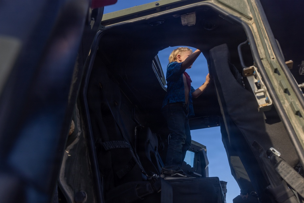 A young blonde child is standing inside what appears to be a military vehicle, likely a HMMWV (Humvee) or similar tactical vehicle, viewed from a low interior angle. The child is reaching upward toward the open door or hatch area, silhouetted against a bright blue sky. The scene suggests a public display or open-house event where civilians can explore military equipment.