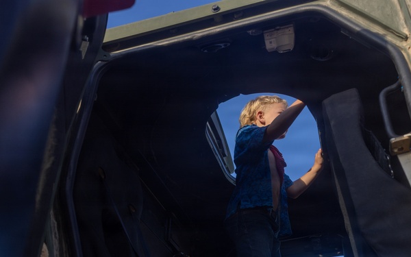 A young blonde child is standing inside what appears to be a military vehicle, likely a HMMWV (Humvee) or similar tactical vehicle, viewed from a low interior angle. The child is reaching upward toward the open door or hatch area, silhouetted against a bright blue sky. The scene suggests a public display or open-house event where civilians can explore military equipment.