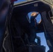 A young blonde child is standing inside what appears to be a military vehicle, likely a HMMWV (Humvee) or similar tactical vehicle, viewed from a low interior angle. The child is reaching upward toward the open door or hatch area, silhouetted against a bright blue sky. The scene suggests a public display or open-house event where civilians can explore military equipment.