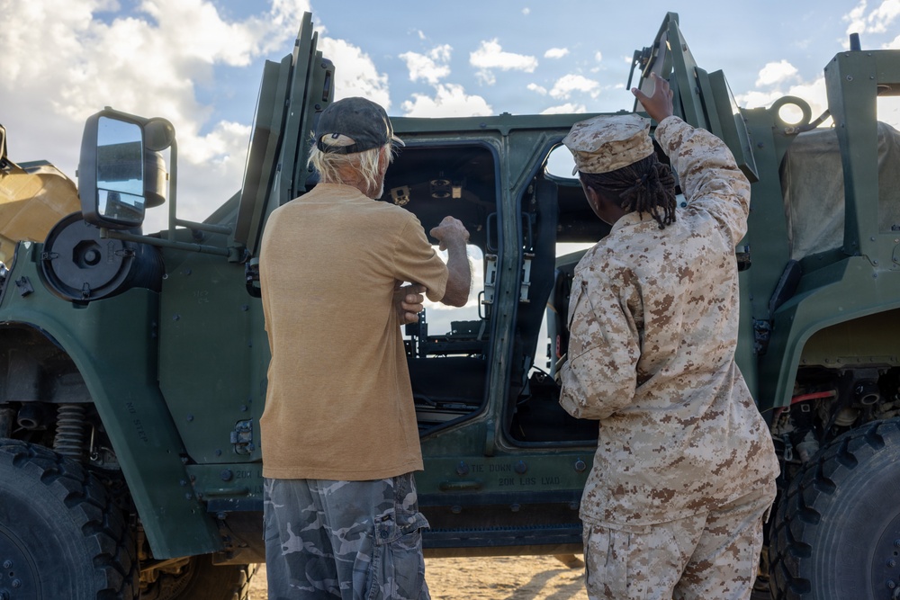Two individuals are standing in front of a large military vehicle with its doors open. One person is wearing US Marine Corps desert MARPAT camouflage uniform, while the other is in civilian clothing. They appear to be inspecting or discussing the vehicle's cab area.