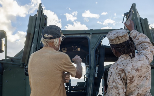 Two individuals are standing in front of a large military vehicle with its doors open. One person is wearing US Marine Corps desert MARPAT camouflage uniform, while the other is in civilian clothing. They appear to be inspecting or discussing the vehicle's cab area.