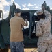 Two individuals are standing in front of a large military vehicle with its doors open. One person is wearing US Marine Corps desert MARPAT camouflage uniform, while the other is in civilian clothing. They appear to be inspecting or discussing the vehicle's cab area.