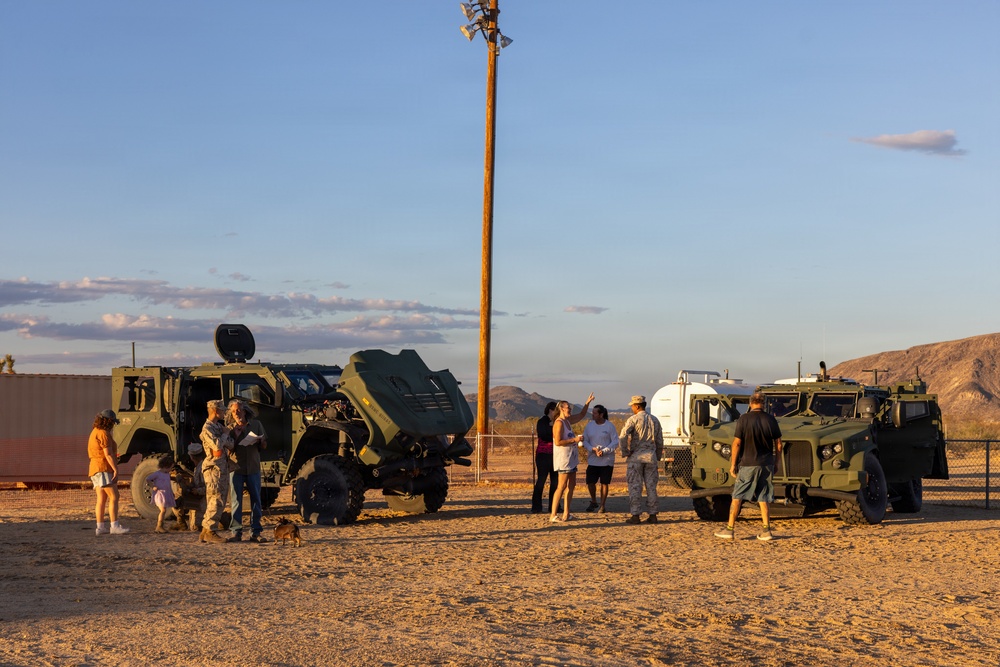 A group of civilians and military personnel are gathered in a desert setting around two olive-green military tactical vehicles, appearing to be an open house or public demonstration event. The scene is photographed during golden hour with warm sunset lighting against a desert landscape with hills in the background. A utility pole with speakers or lights stands prominently in the center of the frame.