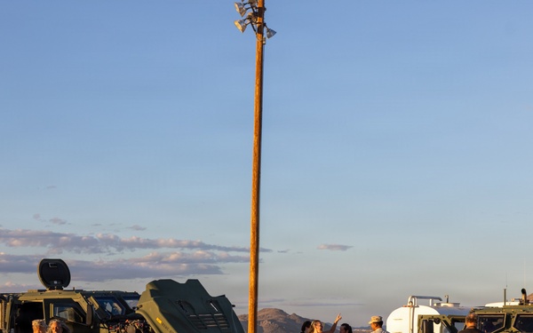 A group of civilians and military personnel are gathered in a desert setting around two olive-green military tactical vehicles, appearing to be an open house or public demonstration event. The scene is photographed during golden hour with warm sunset lighting against a desert landscape with hills in the background. A utility pole with speakers or lights stands prominently in the center of the frame.