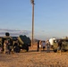 A group of civilians and military personnel are gathered in a desert setting around two olive-green military tactical vehicles, appearing to be an open house or public demonstration event. The scene is photographed during golden hour with warm sunset lighting against a desert landscape with hills in the background. A utility pole with speakers or lights stands prominently in the center of the frame.