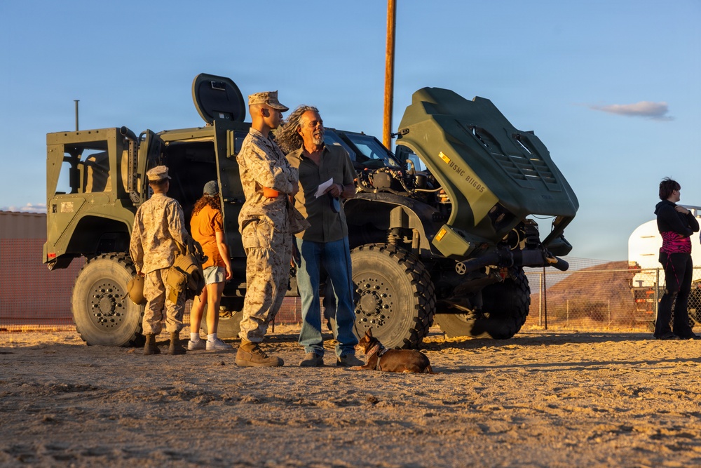 A military open house or community engagement event taking place in an arid outdoor setting, likely a U.S. Marine Corps base. Two Marines in desert camouflage uniforms interact with civilians near military tactical vehicles. The scene is lit by warm golden-hour sunlight, suggesting late afternoon.
