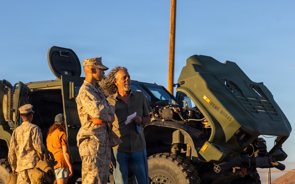 A military open house or community engagement event taking place in an arid outdoor setting, likely a U.S. Marine Corps base. Two Marines in desert camouflage uniforms interact with civilians near military tactical vehicles. The scene is lit by warm golden-hour sunlight, suggesting late afternoon.
