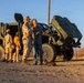 A military open house or community engagement event taking place in an arid outdoor setting, likely a U.S. Marine Corps base. Two Marines in desert camouflage uniforms interact with civilians near military tactical vehicles. The scene is lit by warm golden-hour sunlight, suggesting late afternoon.