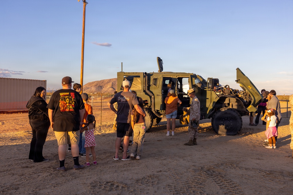 A group of civilians, including children, are gathered around a military tactical vehicle (appears to be a Joint Light Tactical Vehicle or similar JLTV/MRAP-type vehicle) in an arid, desert environment. One individual in military camouflage uniform appears to be conducting a demonstration or tour of the vehicle. The setting suggests a public military open house or community outreach event, likely at or near a military installation.