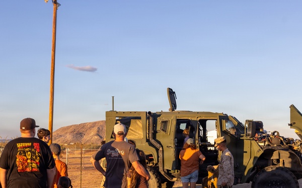 A group of civilians, including children, are gathered around a military tactical vehicle (appears to be a Joint Light Tactical Vehicle or similar JLTV/MRAP-type vehicle) in an arid, desert environment. One individual in military camouflage uniform appears to be conducting a demonstration or tour of the vehicle. The setting suggests a public military open house or community outreach event, likely at or near a military installation.
